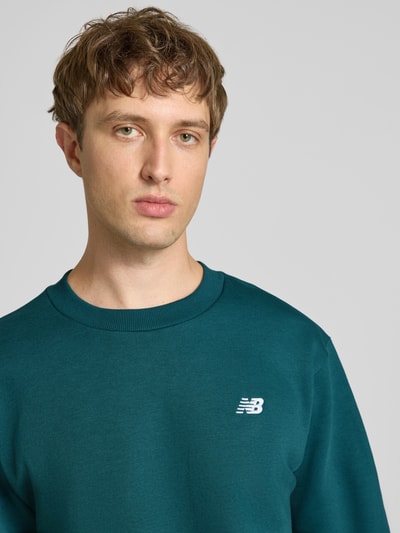 Männermodel trägt Sweatshirt mit Rundhalsausschnitt und Label-Stitching von New Balance in Petrol, Baumwoll-Mix, Nahaufnahme Oberkörper.