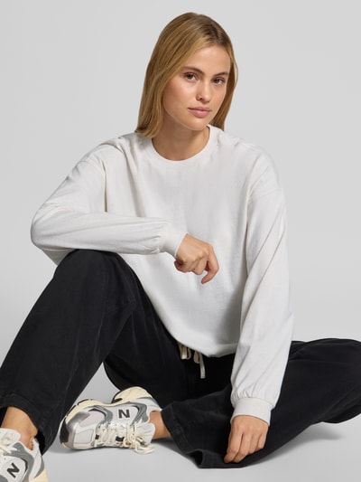 Frauenmodel trägt Relaxed Fit Langarmshirt aus reine Baumwolle von DRYKORN in Weiß mit gerippter Blende und Bündchen, schwarze Hose; Nahaufnahme Oberkörper.
