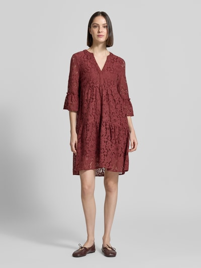 Kobiece modelka nosi sukienkę z koronki z mieszanki bawełny, model 'HONEY' od Vero Moda, jednokolorowy efekt, krój loose fit, długość do kolan, dekolt w serek, koronkowy wzór, rękaw o dł. 3/4 z rozkloszowanymi zakończeniami, wykończenie materiału: koronka, kolor bordowy, widok całej sylwetki z przodu.