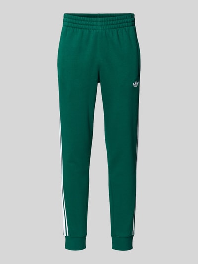 Produktfoto, grüne Sweatpants mit elastischem Bund von adidas Originals aus Baumwoll-Elasthan-Mix, locker geschnitten, weiße Streifen und Label-Stitching.