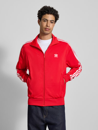 Męski model nosi Sweatjacke mit Stehkragen od adidas Originals, czerwona z białymi paskami, regular fit, widok całej sylwetki z przodu.