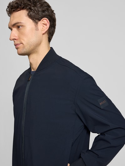 Männermodel trägt Regular Fit Blouson mit Baseball-Kragen Modell 'OSURF' von BOSS Orange in Dunkelblau, Nahaufnahme Oberkörper. Unifarbene Optik mit Reißverschluss, Label Patch, gerippten Bündchen und seitlichen Reißverschlusstaschen.