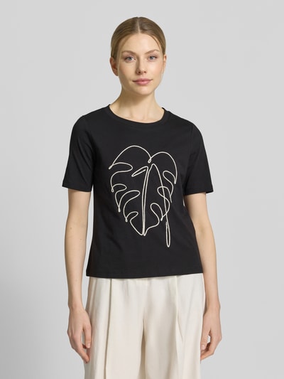 Kobiece model nosi T-Shirt mit Motiv-Stitching od Christian Berg Woman Selection z bawełny, krój regular fit, okrągły dekolt, długość tyłu 54 cm, czarny z białym wyhaftowanym motywem liścia, widok całej sylwetki z przodu.