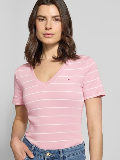 Modelka kobieca nosi Slim Fit T-Shirt aus reiner Baumwolle od Tommy Hilfiger, różowe w paski, krótkie rękawy, logo na piersi, jersey, krój slim fit, dwutonowy wzór, dekolt w serek, widok na górną część ciała z przodu.