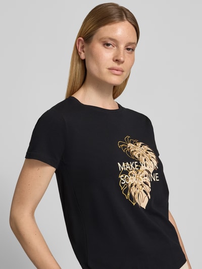 Kobieta modelka nosi damski t-shirt z bawełny, krój regular fit, model 'RINA' od Soyaconcept z nadrukiem z napisem i motywem, okrągły dekolt, długość tyłu w rozmiarze S 60 cm, kolor czarny, faktura gładka. Zbliżenie na górną część ciała.