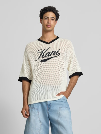 Männermodel trägt T-Shirt in Häkel-Optik mit V-Ausschnitt von KARL KANI, Baumwoll-Mix, Loose-Fit, Label-Stitching, creme mit schwarzen Details, Rückenlänge 68 cm bei Größe M, Ganzkörperansicht von vorne.