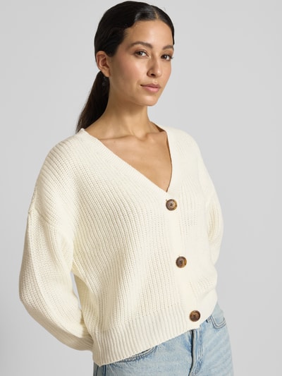 Modelka nosi kardigan rozpinany z dekoltem w serek, krój oversized, model 'LEA' od VERO MODA, kremowa faktura prążkowana, zbliżenie na górną część ciała.