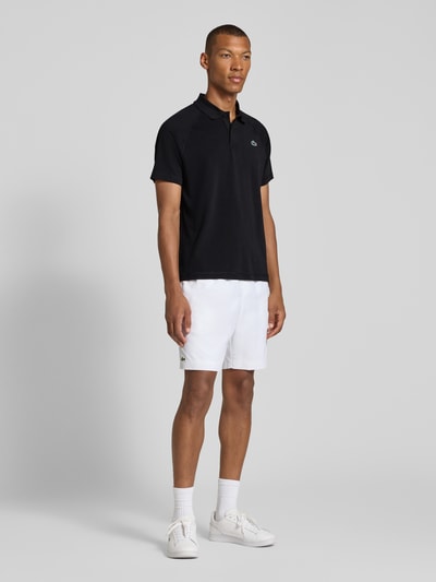 Męskki model nosi czarny Regular Fit Sport-T-Shirt mit Polokragen von Lacoste, białe szorty, sportowe buty, neutralne tło, Ganzkörperansicht von vorne. T-shirt Regular Fit Sport-T-Shirt mit Polokragen od Lacoste Sport.