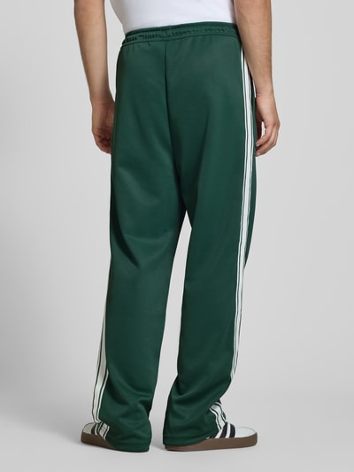 Männermodel trägt Wide Leg Sweatpants mit elastischem Bund von Pegador in Dunkelgrün mit weißen Seitenstreifen, Ganzkörperansicht Rückansicht.