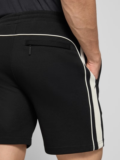 Mężczyzna model nosi czarne szorty z wyhaftowanym logo model 'EGEO' od SERGIO TACCHINI, mieszanka bawełny z dodatkiem streczu, luźny krój, elastyczny pas z trokiem, wpuszczane kieszenie boczne, szerokość w rozmiarze M: 40 cm, długość wewnętrzna nogawki: 20 cm, widok z tyłu na całe nogi.