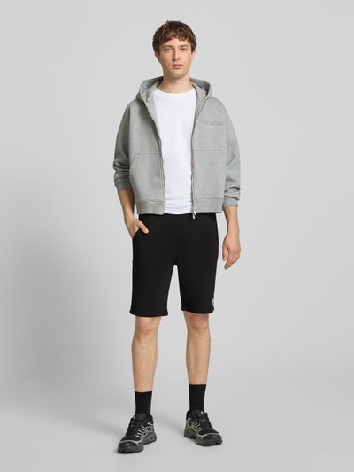 Männermodel trägt Shorts mit Eingrifftaschen und Logo-Print von Alpha Industries aus Baumwoll-Mix in Schwarz, Relaxed-Fit, Ganzkörperansicht von vorne.