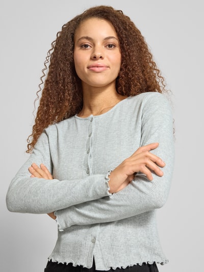 Modelka kobieca ma na sobie Slim Fit Cardigan aus Baumwoll-Viskose-Mix Modell 'MARIE' od Pieces, ażurowy wzór, okrągły dekolt, długi rękaw, melanż, wykończenie frill, listwa guzikowa, mieszanka bawełny i wiskozy. Zbliżenie na górną część ciała z przodu.
