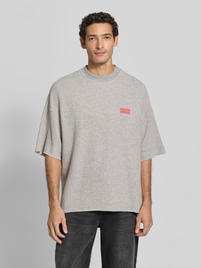 Männermodel trägt Oversized T-Shirt aus Baumwoll-Mix von American Vintage, meliert grau mit rotem Logo-Print, Ganzkörperansicht von vorne. Rückenlänge bei Größe M/L: 70 cm, elastischer Baumwoll-Mix, Oversized Fit, Rundhalsausschnitt.