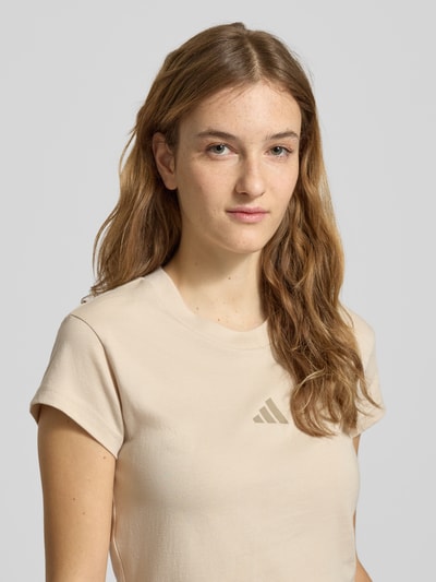 Kobiece model nosi Cropped T-Shirt mit Label-Print von adidas Sportswear w kolorze beżowym, czysta bawełna, krój slim fit, prążkowany okrągły dekolt, nadruk z logo, długość pleców w rozmiarze S: 45 cm. Zbliżenie na górną część ciała.