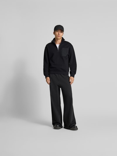 Mannelijk model draagt Troyer aus reiner Baumwolle van MAISON KITSUNÉ in zwart, regular fit, borstzak, label-detail, volledige lichaamsvooraanzicht.