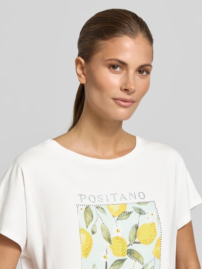 Kobiece modelka nosi T-Shirt mit Motiv-Print und Ziersteinbesatz od Oui, mieszanka bawełny i modalu, krój regular fit, okrągły dekolt, nadruk z motywem cytryn na białym tle, długość tyłu 59 cm w rozmiarze 36, damski t-shirt, zbliżenie na górną część ciała.