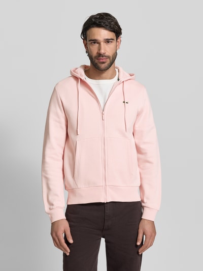 Männermodel trägt Regular Fit Sweatjacke aus Baumwoll-Mix von Lacoste in Hellrosa mit Logo-Patch, Kapuze und Reißverschluss, Ganzkörperansicht von vorne.