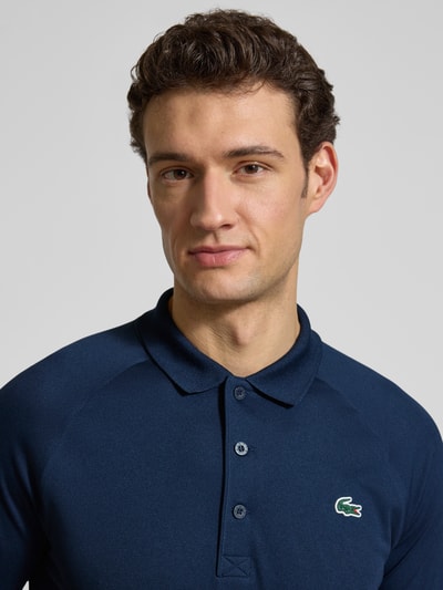 Męski model nosi Regular Fit Sport Poloshirt mit Label Patch od Lacoste, efekt: jednolity kolor, krój regular fit, naszywka z logo, kołnierzyk polo, zapięcie na 3 guziki, krótki rękaw, wykończenie materiału: pika; zbliżenie na górną część ciała.