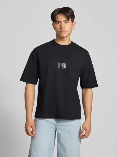 Männermodel trägt T-Shirt mit Logo-Print und Rundhalsausschnitt von REVIEW in Schwarz, Reine Baumwolle, Loose-Fit, Ganzkörperansicht von vorne. Rückenlänge bei Größe M: 69 cm.