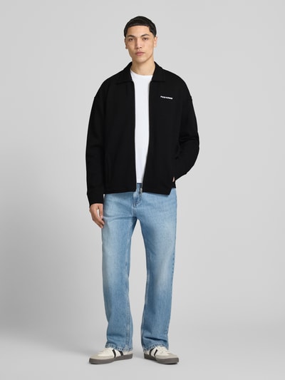 Männermodel trägt Oversized Sweatjacke mit Strukturmuster Modell 'Tate' von Pegador in Schwarz, Baumwoll-Mix mit Stretch-Anteil, Umlegekragen, Reißverschluss, Label-Stitching, kombiniert mit heller Jeans und weißen Sneakern, Ganzkörperansicht von vorne. Kontext: Herren Sweatjacke, Rückenlänge bei Größe M 69 cm, Ärmellänge bei Größe M 73 cm.