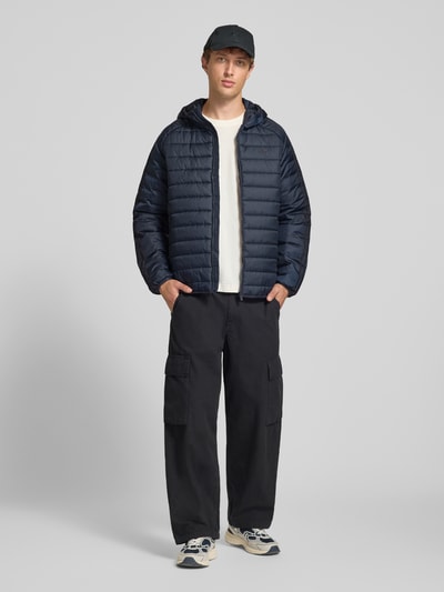 Männermodel trägt Regular Fit Steppjacke mit Reißverschluss von adidas Originals in Dunkelblau, dezente Struktur, Ganzkörperansicht von vorne.