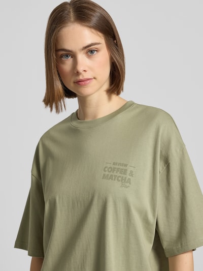Kobiece modelka nosi Oversized T-Shirt mit Statement-Print od REVIEW, czysta bawełna, krój oversize, okrągły dekolt, nadruk z przodu, długość tyłu w rozmiarze S: 70 cm, jasna oliwkowa zieleń, gładka tekstura, prosty fason, zbliżenie na górną część ciała.