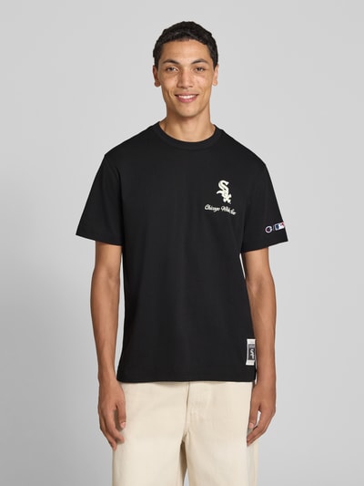 Męski model trägt Relaxed Fit T-Shirt aus reiner bawełna von Champion, czarny z nadrukiem z napisem, wyhaftowane logo, Ganzkörperansicht von vorne, długość tyłu w rozmiarze M: 70 cm.