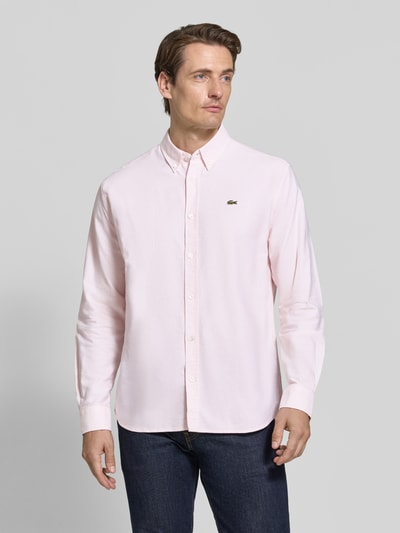 Männermodel trägt Regular Fit Freizeithemd aus reiner Baumwolle von Lacoste in Hellrosa mit Knopfleiste und Logo, Ganzkörperansicht von vorne.