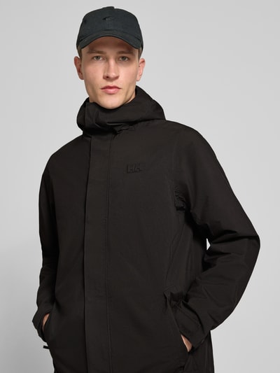 Männermodel trägt Regular Fit Mantel mit Kapuze Modell 'Brussel' von Helly Hansen, schwarz, Logo-Detail, Nahaufnahme Oberkörper. Rückenlänge 96 cm, Schulterbreite 46 cm, Brustweite 51 cm, Ärmellänge 66 cm.