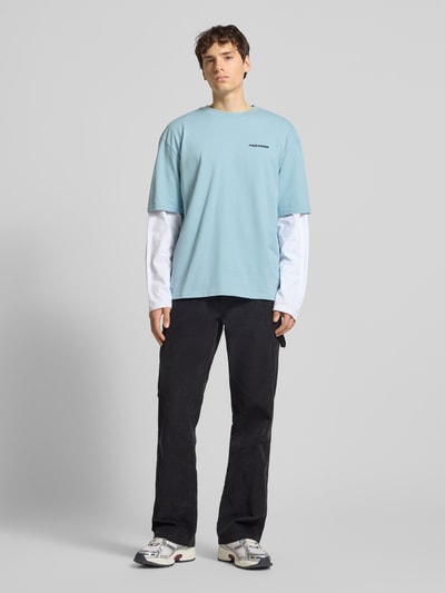 Männermodel trägt Oversized T-Shirt mit Label-Stitching von Pegador in Hellblau, reine Baumwolle, gerippter Rundhalsausschnitt, Ganzkörperansicht von vorne.