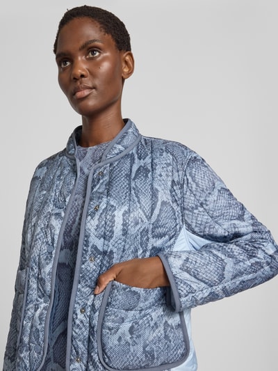 Frauenmodel trägt Regular Fit Jacke in Stepp-Optik mit Stehkragen von Marc Cain, Schlangen-Print in Blau, Nahaufnahme Oberkörper, Allover-Muster, durchgängige Knopfleiste, aufgesetzte Taschen, glatte und strukturierte Texturen, Kontext: Rückenlänge 56 cm, Schulterbreite 43 cm, Brustweite 50 cm, Ärmellänge 60 cm.