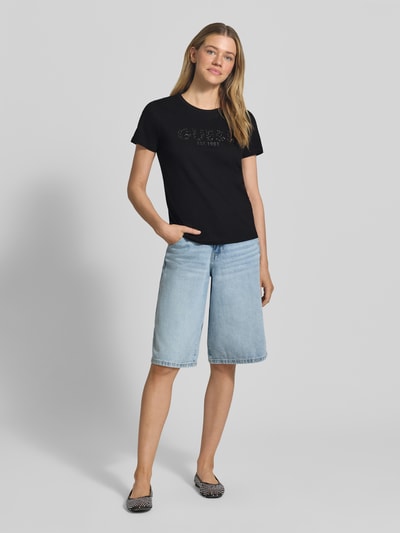 Frauenmodel trägt T-Shirt mit Label-Print und Strasssteinbesatz von Guess aus schwarzer Baumwolle, Straight Fit, Ganzkörperansicht von vorne.