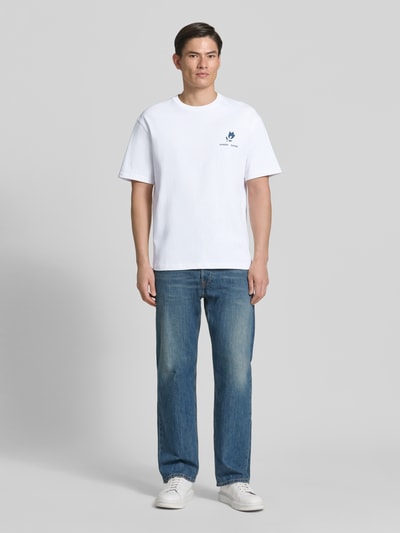Męski model nosi T-Shirt mit Label-Print Modell 'COHEN' od Samsøe Samsøe z czystej bawełny organicznej, krój relaxed fit, biały kolor, nadruk z przodu, okrągły dekolt, długość tyłu w rozmiarze M: 71 cm, widok całej sylwetki z przodu.