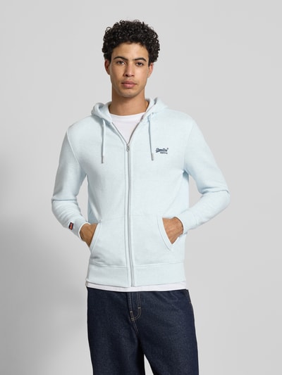 Męski model nosi Regular Fit Sweatjacke mit Logo-Stitching od Superdry z mieszanki bawełny, błękitna, zamek błyskawiczny, kieszeń kangurka, kaptur z tunelem, wykończenia elastyczne, długość tyłu 69 cm, długość rękawów 65 cm, widok całej sylwetki z przodu.