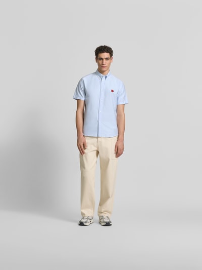 Männermodel trägt Slim Fit Freizeithemd mit Logo-Stitching von Kenzo aus reiner Baumwolle, Button-Down-Kragen, Kurzarm, hellblau, beige Hose, Ganzkörperansicht von vorne.