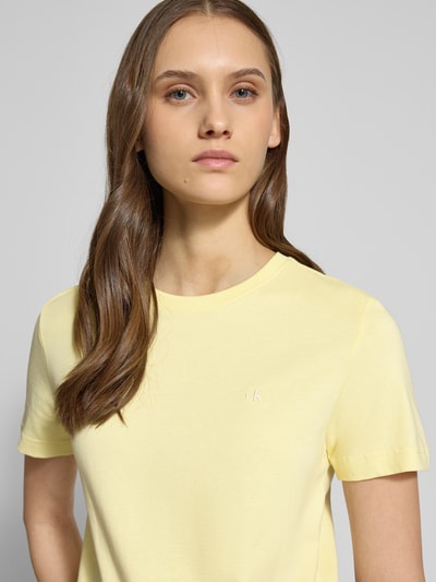 Kobiece model prezentuje T-shirt mit Rundhalsausschnitt und Label-Stitching von Calvin Klein Jeans z bawełny, krój regular fit, okrągły dekolt, wyhaftowane logo, długość tyłu w rozmiarze S: 52 cm, jasny żółty kolor, gładka faktura, widok na górną część sylwetki, z przodu.