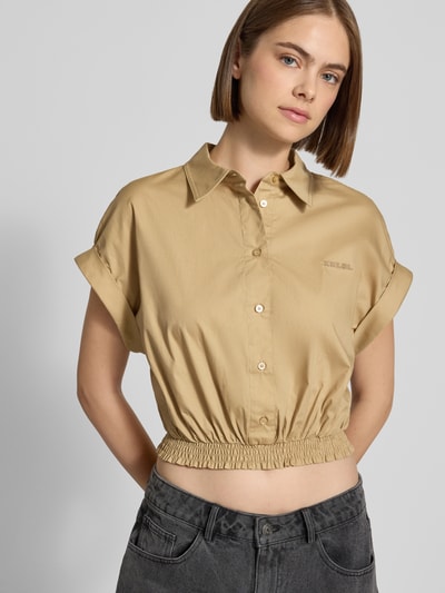 Modelka nosi Cropped Hemdbluse aus reiner Baumwolle mit Logo-Stitching von Karo Kauer w kolorze beżowym, krój krótki, wyhaftowane logo, kołnierzyk koszulowy, krótki rękaw, z listwą guzikową i elastycznym pasem, długość tyłu w rozmiarze S: 45 cm, widok na górną część sylwetki z przodu.