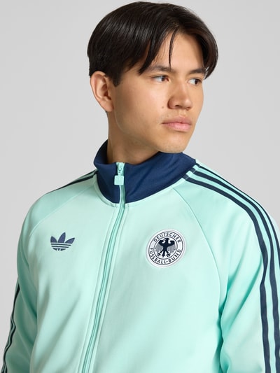 Männermodel trägt Regular Fit Trainingsjacke mit Baumwoll-Anteil und DFB-Logo von adidas Originals, Optik: Unifarben, Nahaufnahme Oberkörper.