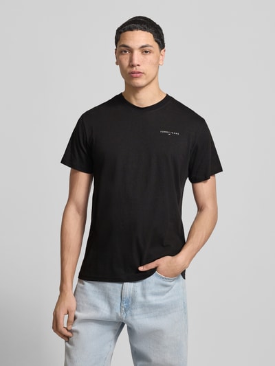 Männermodel trägt Herren T-Shirt Slim Fit T-Shirt aus reiner Baumwolle Modell 'LINEAR CHEST TEE' von Tommy Jeans, Optik: Unifarben, Passform: Regular-Fit, Applikation: Label-Print, Ausschnitt: Rundhals, Ärmelform: Kurzarm, Material: Reine Baumwolle, schwarze Farbe, glatte Textur, Ganzkörperansicht von vorne.