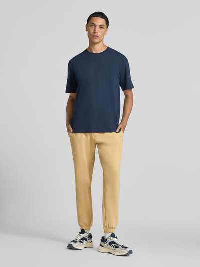 Männermodel trägt Slim Fit Sweathose aus Baumwoll-Mix von Tommy Jeans in Beige, glatte Oberflächen, lockere Passform, Ganzkörperansicht von vorne. Herren Sweathose von Tommy Jeans.