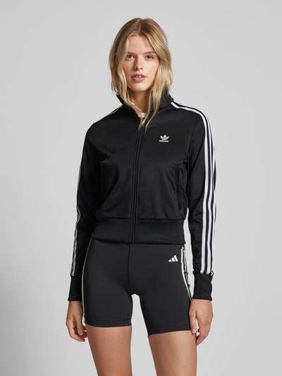 Kobiece modelka nosi czarną damską adidas Originals Trainingsjacke mit Logo-Stitching Modell 'FIREBIRD', proste kolory, białe paski, widok całej sylwetki z przodu.