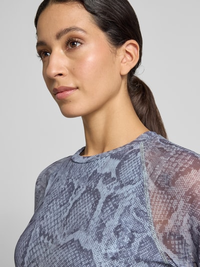 Frauenmodel trägt Longsleeve aus Mesh mit Schlangen-Muster von Marc Cain in Blau- und Grautönen, semitransparente Ärmel, Nahaufnahme Oberkörper.