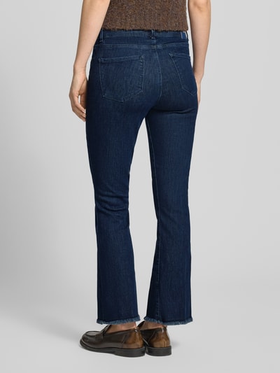Frauenmodel trägt Bootcut Jeans mit Fransen Modell 'SHAKIRA' von Brax aus Baumwoll-Mix mit Stretch-Anteil, Gesäßtaschen, verkürzte Passform, dunkelblaue Jeans-Optik, Fransen-Saum, Ganzkörperansicht Rückansicht.