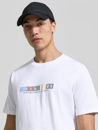 Männermodel trägt T-Shirt aus Bio-Baumwolle Modell 'NANTUCKET' von Jack & Jones, weiß mit buntem Label-Print, Nahaufnahme Oberkörper.