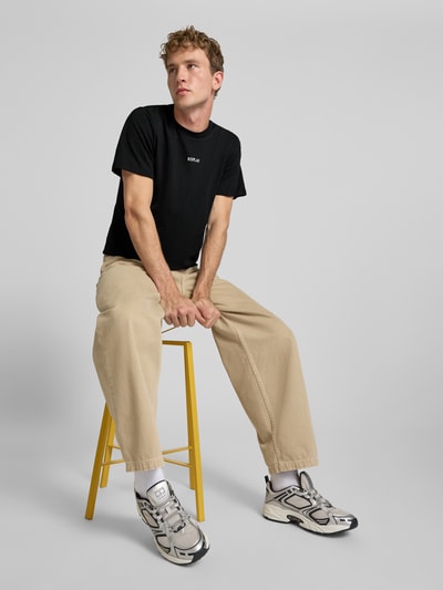 Männermodel trägt T-Shirt mit Rundhalsausschnitt und Logo von Replay, schwarze Baumwolle, Slim Fit, bequeme beige Hose, Ganzkörperansicht von vorne.