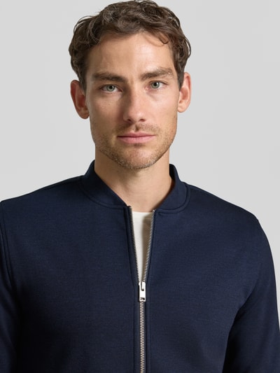 Męskamodel nosi Slim Fit Sweatjacke mit Stehkragen od Lindbergh, Mieszanka materiałów z dodatkiem streczu, zamek błyskawiczny, wpuszczane kieszenie, długość tyłu 67 cm, długość rękawów 66 cm, krój slim fit, kolor granatowy, tekstura gładka, zbliżenie na górę ciała.