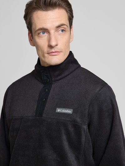 Männermodel trägt Fleecepullover mit Stehkragen und Label-Detail von Columbia, schwarze Farbe, Knopfleiste, Nahaufnahme Oberkörper. Herren Regular-Fit Modell STEENS MOUNTAIN, Rückenlänge 70 cm, Ärmellänge 66 cm.