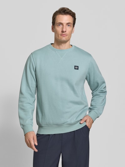 Männermodel trägt Regular Fit Sweatshirt aus Baumwoll-Mix von LERROS in hellem Blaugrün mit Logo-Patch und gerippten Abschlüssen, Ganzkörperansicht von vorne.