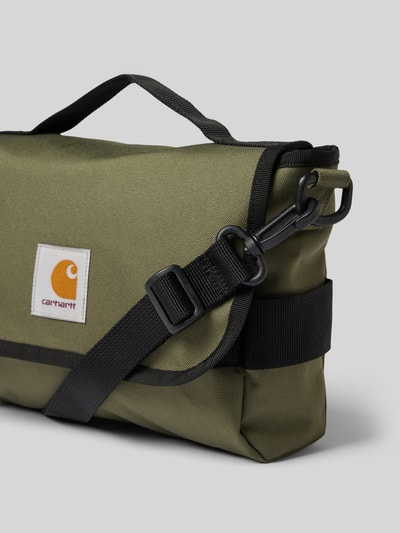 Produktfoto, Tasche mit Logo-Patch von Carhartt Work In Progress aus olivgrünem, wasserabweisendem Stoff mit schwarzem Schulterriemen, Detailaufnahme. Hauptfach mit Überschlag, abnehmbarer Schulterriemen, Label vorne, Maße circa 28x19x6 cm.