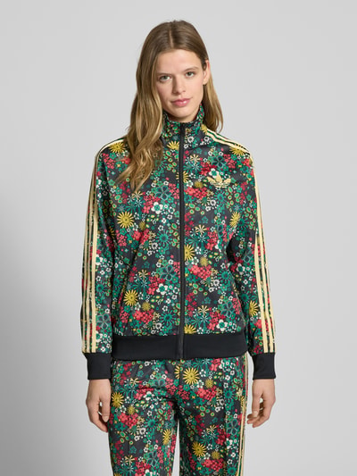 Vrouwenmodel draagt Regular Fit Jacke mit Logo-Stitching Modell 'Firebird' van adidas Originals met florale allover-print, staande kraag, steekzakken, elastische boorden en ritssluiting, kleurrijke bloemen op donkere achtergrond, vooraanzicht bovenlichaam.
